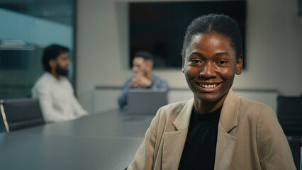 Webcam view African American ethnic woman business coach businesswoman smiling toothy video call conference chat looking at camera sitting at table on office on background of talking men colleagues
