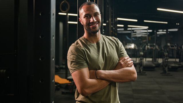 Smiling happy African American fitness trainer posing with crossed arms in gym. Latino guy sportsman male strong muscular athlete bodybuilder man smile at camera in sport club after fitness workout