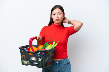 Young Asian woman holding a shopping basket full of food isolated on white background having doubts