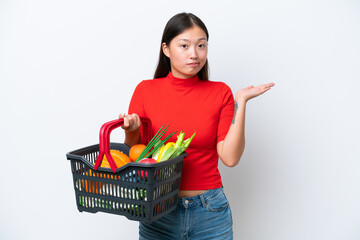 Obraz na płótnie Canvas Young Asian woman holding a shopping basket full of food isolated on white background having doubts while raising hands