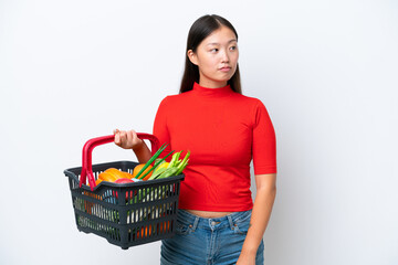 Obraz na płótnie Canvas Young Asian woman holding a shopping basket full of food isolated on white background looking to the side