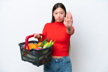 Obraz na płótnie Canvas Young Asian woman holding a shopping basket full of food isolated on white background making stop gesture