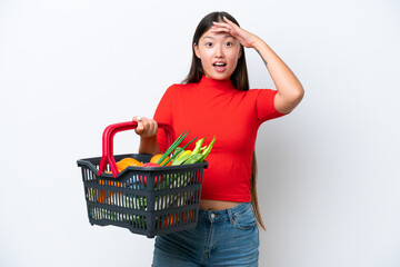 Young Asian woman holding a shopping basket full of food isolated on white background doing surprise gesture while looking to the side