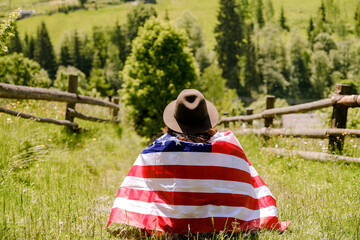 A woman sits on a hill covered with an American flag and looks into the distance in the summer. USA Independence Day celebration.