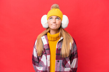 Young beautiful woman wearing winter muffs isolated on red background and looking up