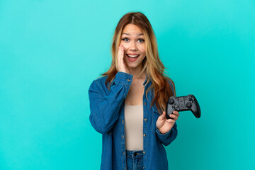 Teenager blonde girl playing with a video game controller over isolated wall with surprise and...