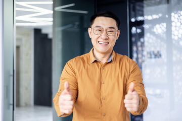 Portrait of successful asian man inside office near window, boss in glasses and shirt looking at camera and showing thumbs up.