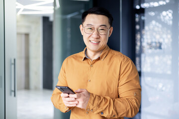Portrait of a successful Asian man inside the office, a man near window smiling and looking at the camera, a businessman holds a phone in his hands, types a message and browses the Internet.