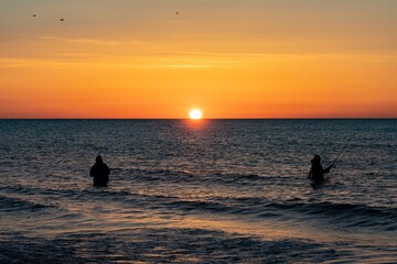 Fototapeta premium Die Silhouetten von zwei Anglern in wasserdichten Hosen angelen vor einem traumhaften orangen Sonnenuntergang in der Ostsee. Sie stehen mitten im Wasser der Ostsee.
