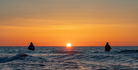 Die Silhouetten von zwei  Anglern in wasserdichten Hosen angelen vor einem traumhaften orangen...