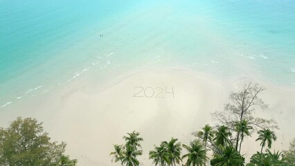 Stunning aerial view of the year 2024 written on the white sandy beach in tropical paradise scenery with palms, crystal clear turquoise water, and a couple leisurely strolling through the shallow sea.