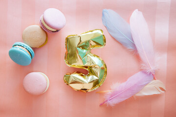 Celebration card with golden air balloon of number five on pink background with feathers and macaroons, anniversary concept