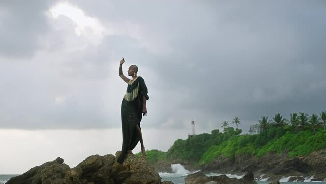 LGBTQIA black person in boutique dress, jewelry stands on rock on stunning ocean coastline skyline. Gender fluid ethnic divine model in luxury outfit fluttering in the wind against dramatic sky.
