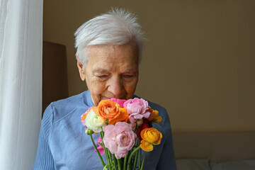 Portrait of happy elderly woman receiving a bouquet of colorful ranunculus flowers and inhaling the sweet aroma. Senior lady sniffing buttercups. Close up, copy space, background.