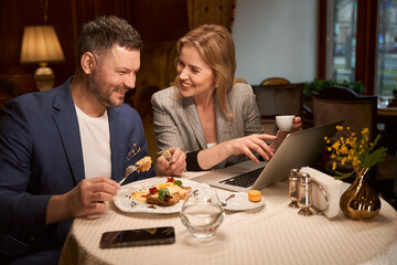 Spouses smiling looking at photos from vacation on laptop at restaurant