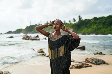 Epatage lgbtq black male posing with hands up on camera on scenic ocean beach. Non-binary ethnic fashion model in long posh dress wears jewellery stands gracefully on sea shore and lighthouse.