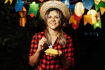 Woman eating Pamonha, a traditional corn-based dessert, at the Festa Junina (June Festival) embracing the flavors and traditions of Brazil's June celebration. - obrazy, fototapety, plakaty