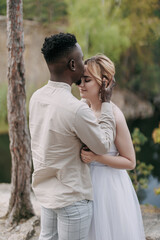 Happy interracial couple newlyweds stands, embraces and kisses against background of lake and forest.