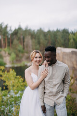 Happy interracial couple newlyweds stands and laughs cheerfully against background of lake and forest.