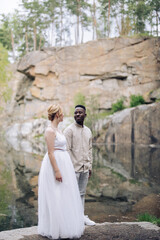 Happy interracial couple newlyweds stands on rock and looks each other against background of lake and canyon.