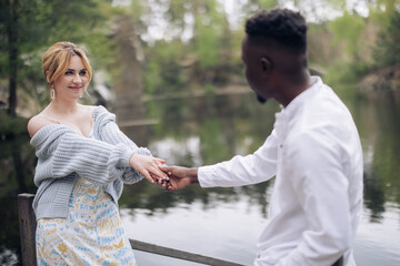 Happy interracial couple holds hands and looks each other and smiles against background of lake.