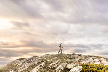 Woman walking with sticks on the mountain at sunset at the top, with colours in the clouds and sun streaks, rocky ground with green bushes, yellow jacket and pink pants.