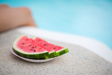 plate with sliced pieces of watermelon on side of outdoor pool. man girl in background