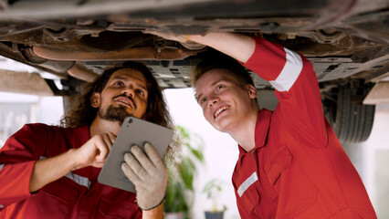two men who are mechanics, working in large garage, two mechanics checking undercarriage car raised high see lower part clearly, holding car check book talk each other before fixing car.