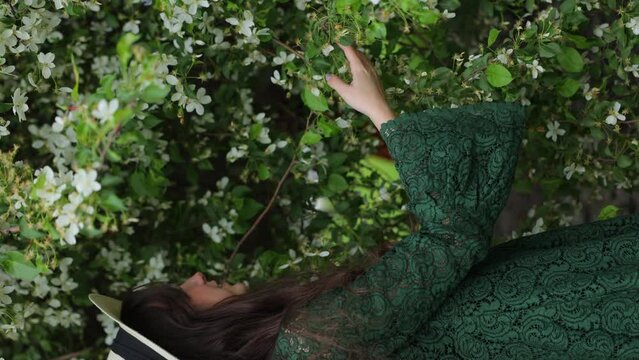 Young woman in a green lace dress and hat is enjoying the blossoms of the apple trees.  Vertical video