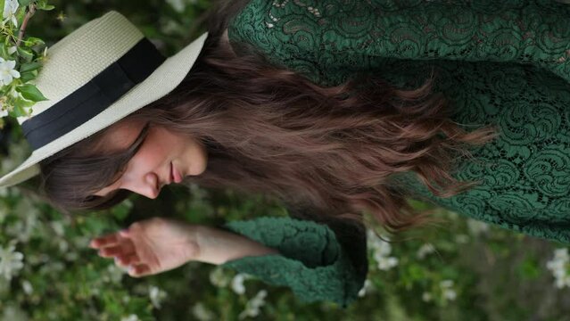 Girl in a green lace dress and hat is playing and fooling around against a backdrop of blossoming apple trees in springtime. The concept of ecology and friendliness to nature. Vertical Video