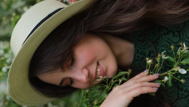 Young pretty woman enjoys standing near flowering spring tree. A girl wearing hat and green dress smiles among blooming apple trees. Spring season concept. Vertical video