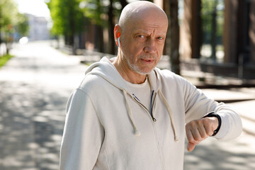 Portrait of the Sporty Mature Jogger Man Standing in the Street Listening to Music on the Earphones, Wearing a Smartwatch and Using it to Calculate Calories. Sport and Healthy Lifestyle Concept