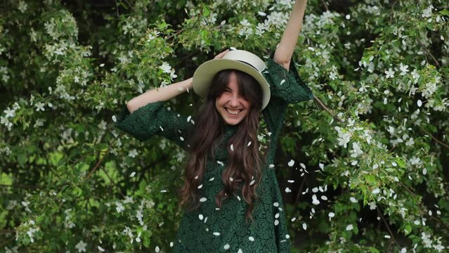 beautiful and joyful brunette woman stands in front of a blossoming apple tree and shakes it. Falling white petals from the blossoming fruit trees. Joy and happiness. The concept of spring and blossom