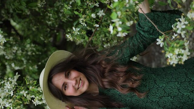 A cheerful and friendly young woman peeks out from behind the branches of a blossoming apple tree. Joy and relaxation in the parks. Vertical video