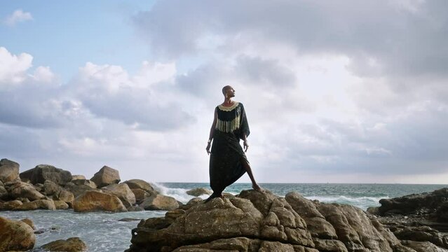 Queer black person gracefully poses in posh dress, handmade jewelry stands on scenic sea cliff top above dramatic cloudy sky, ocean waves. Lgbtq biethnic gay model in brass accessories.