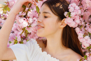 Obraz na płótnie Canvas Portrait of beautiful young woman with sincere smile on face posing with closed near blooming sakura tree. Cute caucasian lady with long brown hair looking at camera
