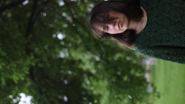 A beautiful and joyful brunette woman in a green dress walks in front of a green garden, with the petals of blossoming fruit trees falling in the background. Vertical video