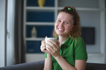 Happy young woman drinking a cup of tea. Dreaming girl sitting in living room with cup of hot coffee enjoying. Pretty woman home.