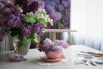 Romantic background with a bouquet of lilacs in a cup, lilac flowers and an open book, scattered paints on a white table.