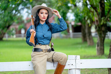Portrait of pretty Asian woman with cowboy style hold rifle gun on her shoulder and sit on fence in...