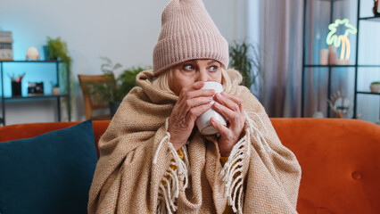 Sick senior grandmother woman in plaid shivering from cold on sofa drinking hot tea in unheated apartment without heating due debt. Unhealthy ill mature pensioner feeling discomfort try to warming up