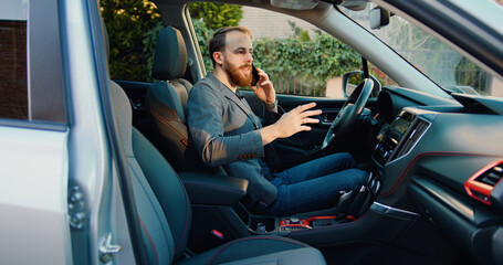 Attractive businessman young male in auto speaking on mobile phone and smiling.. Cheerful casual man sitting in luxury car and chatting on smartphone.