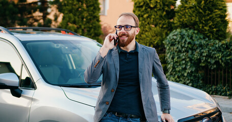 Stylish businessman talking on cell phone answering business call when leans on her modern car...