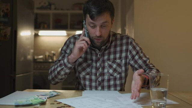 Man argues over phone after getting utility bill in evening at home sitting at table in kitchen. Male swearing on phone looking at bank receipt calculating taxes, examining an invoice document. 