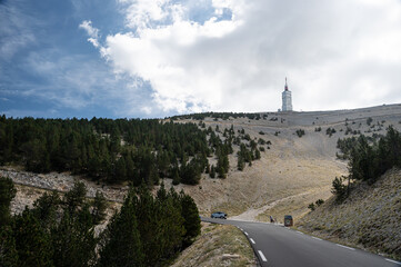 View of the Mont Ventoux, Ventoux mountain named Le Géant de Provence. In the foreground a road, slopes covered with stones and pine trees. A cloudy sky. 