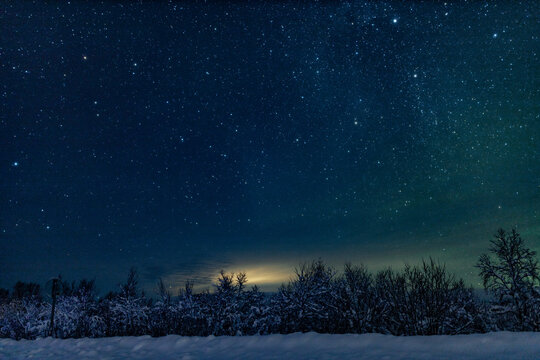 Landscape of the night sky with bright stars and green northern lights on the background of the road, trees, nature, snow in winter. Wide angle