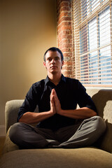 Young Caucasian man sitting on sofa meditating.