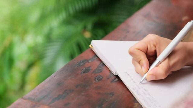 closeup of a hand journaling in the bottom right corner of the picture on a table that cuts the clip diagonally in front of blurry green jungle
