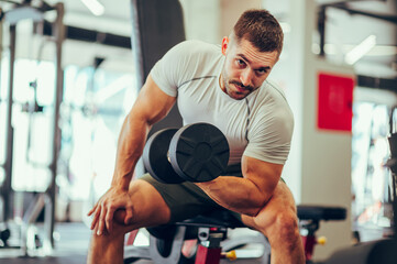 A young strong man is doing exercises for his biceps with heavy weights while making eye contact in a gym.