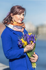 A young woman in a coat stands with a bouquet on the embankment in the park. The concept of the spring holiday is March 8, Easter, Women's Day.